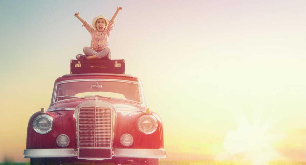 Young kid sitting on top of an old car at sunset with hands in the air